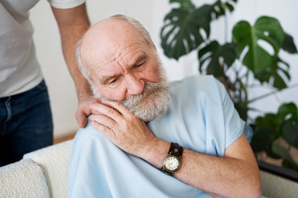 Caregiver rests hand on shoulder of seated elderly man, comforting.